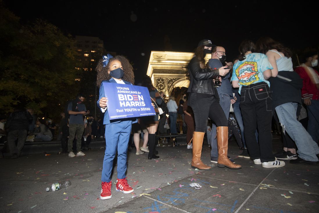 A young Black girl holds a sign that says Young Americans for Biden Haris with the Washington Square Park arch behind her